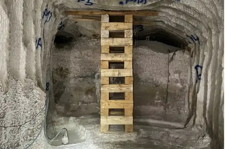 Foto eines Bergwerks unter Tage. Boden, Decke und Wände bestehen aus grauem Salzgestein. Eine Stütze aus Holz stützt die Decke. Im Hintergrund befinden sich Hohlräume an der Decke. Links unten am Boden ist eine kleine Pfütze mit Salzwasser.