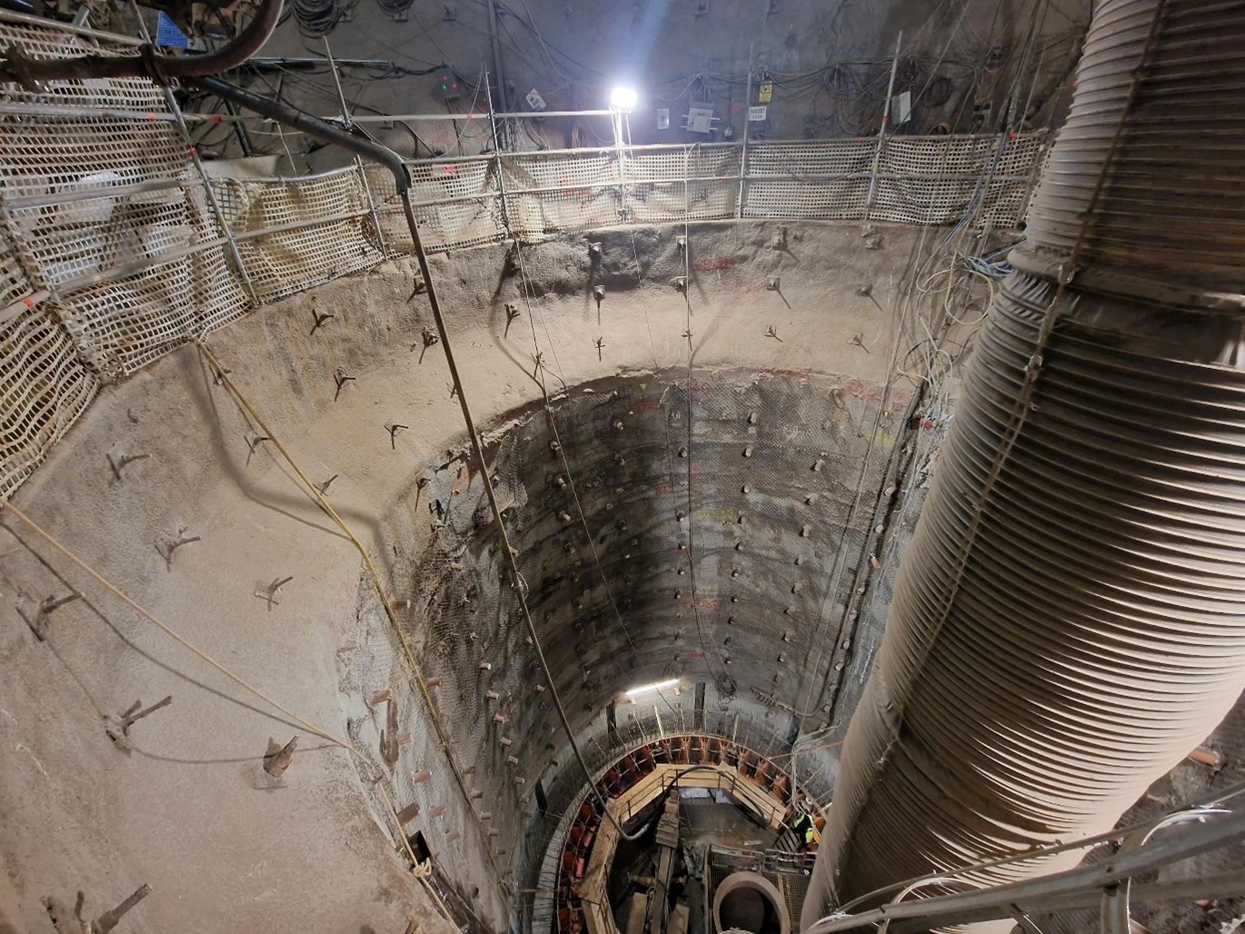 Das Foto von unter Tage im Endlager Konrad zeigt ein senkrecht nach unten führenden Schacht, der mit Gitterdraht und dicken Metallstäben gesichert ist. Von den Metallstäben ragen nur die Köpfe aus dem Gestein. Rechts im Bild ist ein großer Schlauch zu sehen.