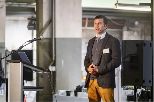 Ein Mann mit kurzen dunklen Haaren, einer dunkelgrauen Anzugjacke, Pullover und Hemd mit Krawatte steht in einer Industriehalle und spricht in ein Headset-Mikrofon. Zwei Schritte vor ihm steht ein Rednerpult mit geöffnetem Laptop.