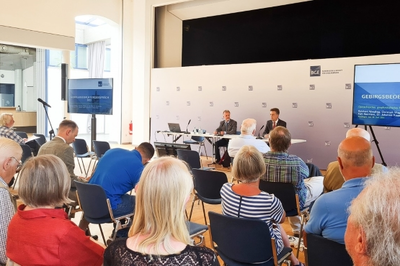 Das Foto zeigt denn Blick in den Veranstaltungsraum. Personen sitzen in mehreren Reihen auf Stühlen und blicken in Richtung Podium. Am Podium sitzen BGE-Mitarbeiter und halten einen Vortrag. Auf zwei Monitoren werden die Präsentationen abgebildet.