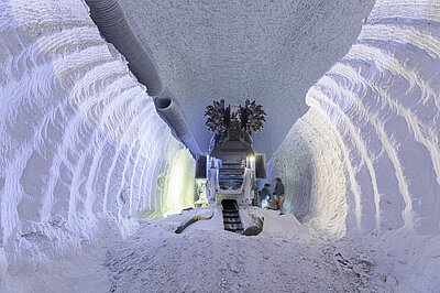  Eine Teilschnittmaschine steht in einer Strecke unter Tage, die durch das Salz gefräst wurde. Die Wände und der Boden bestehen aus schneeweißem Salz. Zwei Personen stehen neben der Maschine.