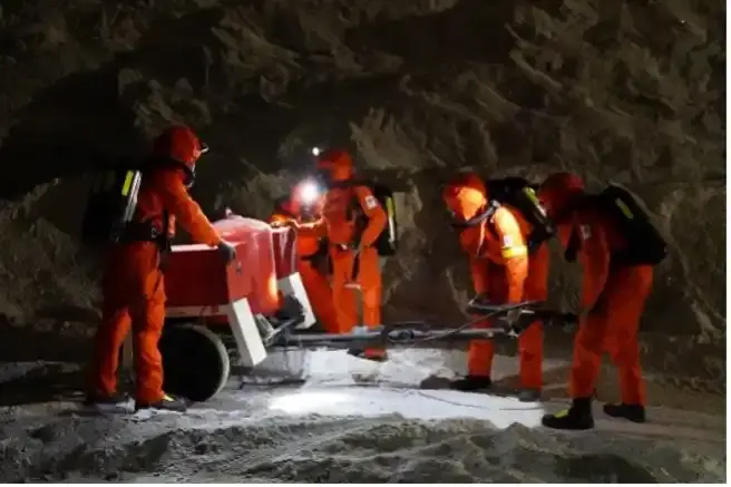 Vier Personen in orangefarbenen Rettungsanzügen bewegen unter Tage in einem Salzbergwerk einen Anhänger.