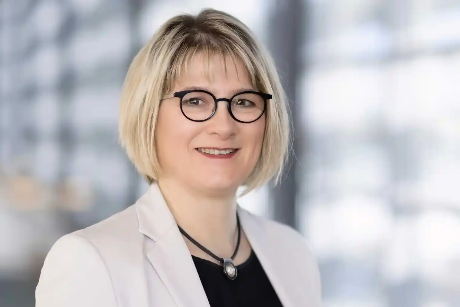 Portrait of a women with a blonde bob and a black pair of glasses is smiling towards the camera.