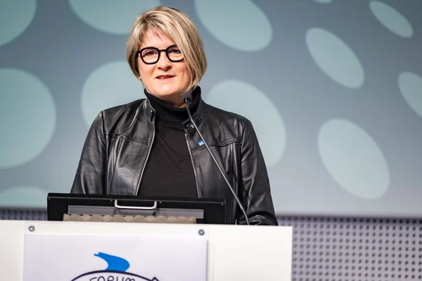 Das Foto zeigt eine Frau mit blondem Bob und dunkler Kleidung, die an einem Podium steht und Richtung Publikum blickt.