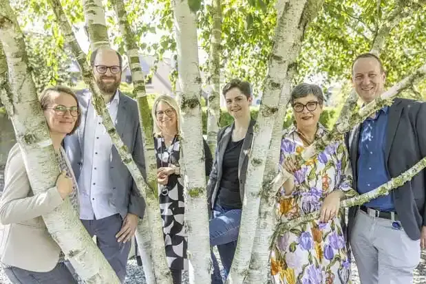 Bei frühlingshaftem Wetter hat sich das Team der Öffentlichkeitsarbeit Standortauswahl zwischen den Ästen einer Birke platziert. Es besteht aus vier Frauen und zwei Männern, die alle freundlich in die Kamera lächeln. 