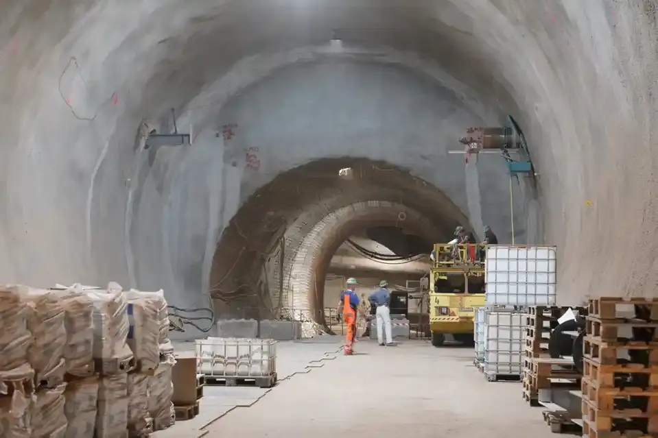 Foto einer tunnelähnlichen Strecke unter Tage. Die Wände sind weißgrau, an den Seiten stehen Paletten und Tanks. In der Mitte des Bildes befinden sich in etwas Entfernung zwei Personen und ein gelbes Baustellenfahrzeug.