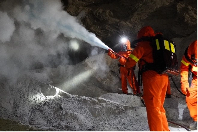 Drei Personen in orangefarbenen Rettungsanzügen befinden sich in einem Salzbergwerk unter Tage. Eine Person hält einen Schlauch und versprüht mit diesem eine Art Nebel.