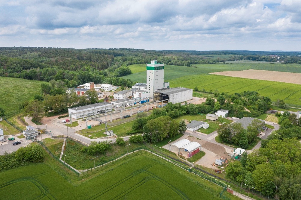 Foto aus der Vogelperspektive eines Industriekomplexes mit mehreren weißen Gebäuden in einer Agrarlandschaft mit Feldern und Waldgebieten.