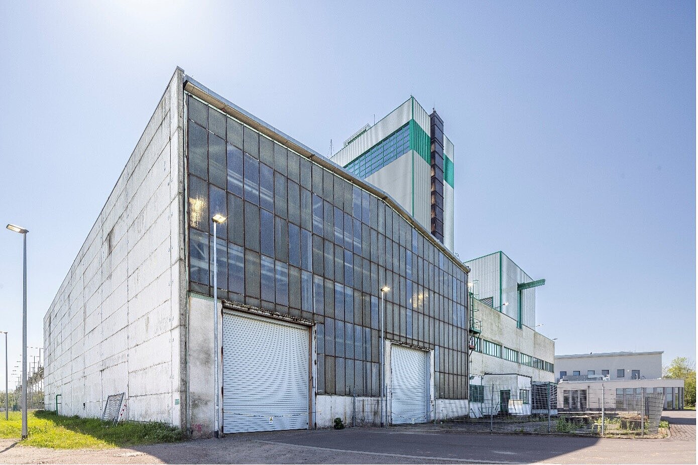 Das Bild zeigt die Fassade einer Halle mit zwei großen Rolltoren. Ein Teil des Gebäudes ist eingezäunt. Hinter der Halle steht ein grün-weißer Turm. 