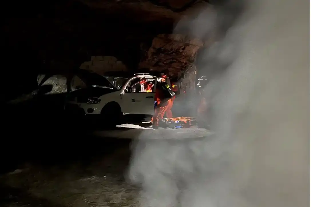 Vier Personen in orangefarbener Schutzkleidung stehen vor einem weißen Auto mit offener Motorhaube. Rechts im Bild ist eine dichte Rauchwolke. 