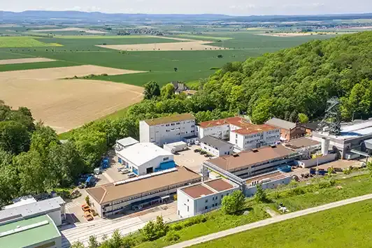 Das Bild zeigt mehrere zwei- und dreistöckige Gebäude sowie Industrieanlagen mit einem Fördergerüst. Die Gebäude sind umgeben von grünem Wald. Den Hintergrund bestimmen Felder und der Horizont mit blauem Himmel. 