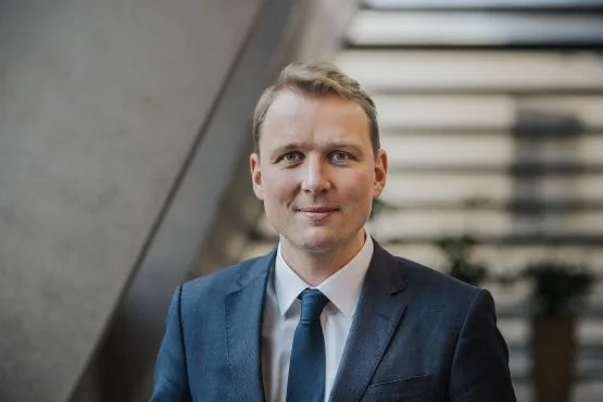 A photo of a blonde man with short hair wearing a dark blue suit with a matching tie. He is smiling towards the camera.