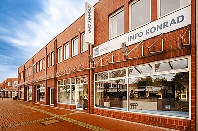 Außendarstellung eines Gebäudes mit roter Backsteinfassade und großen Ladenfenstern in einer Fußgängerzone. Über dem Eingang hängt ein Schild mit der Aufschrift „Info Konrad“.