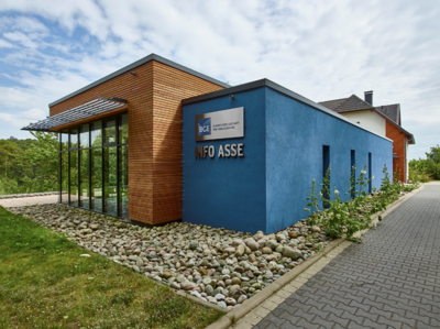 Foto eines Gebäudes mit flachem Dach. Ein Teil des Gebäudes ist mit Holz verkleidet, der andere Teil ist blau gestrichen. Auf dem Gebäude befindet sich der Schriftzug „Info Asse“.