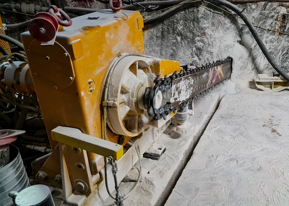 Das Bild zeigt eine Maschine mit einem gelben Motorgehäuse, an der ein etwa zwei Meter langes Sägeblatt montiert ist. Die gesamte Anlage steht auf einem geraden Boden aus (Spezial-)Beton. In den Boden vor der Säge ist bereits ein tiefer Schlitz gesägt. Das Sägeblatt sägt gerade einen weiteren Schlitz in die Wand aus Salz.