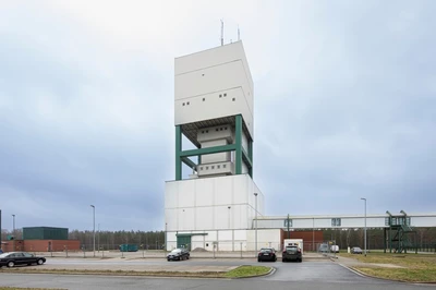 Ein weißes Industriegebäude erhebt sich in den bewölkten Himmel. Vor dem Gebäude befindet sich ein asphaltierter Parkplatz mit wenigen Autos.