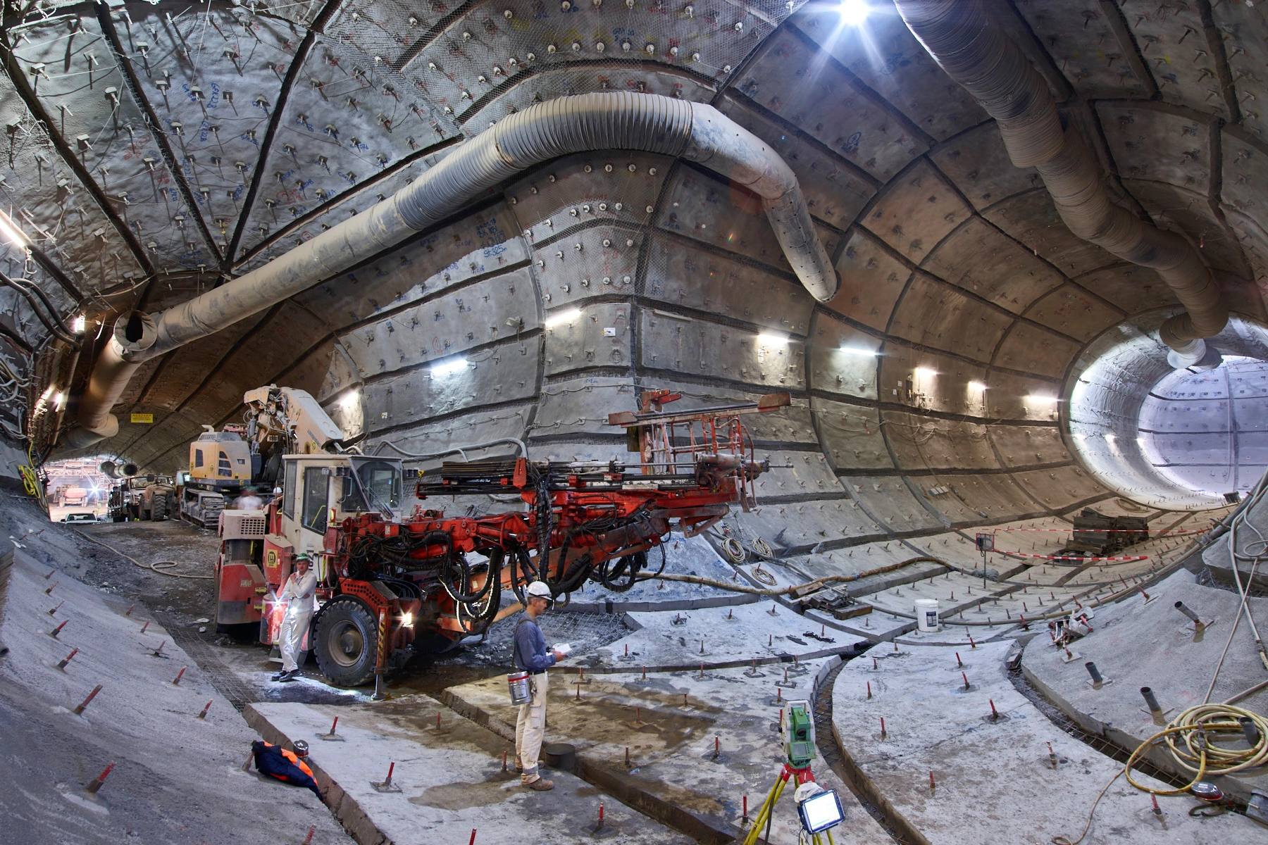 Große Maschinen stehen in einer ausgebauten Strecke im Bergwerk. Aus den Wänden ragen unzählige Gebirgsanker. Im Vordergrund stehen zwei Bergleute in Arbeitskleidung. 