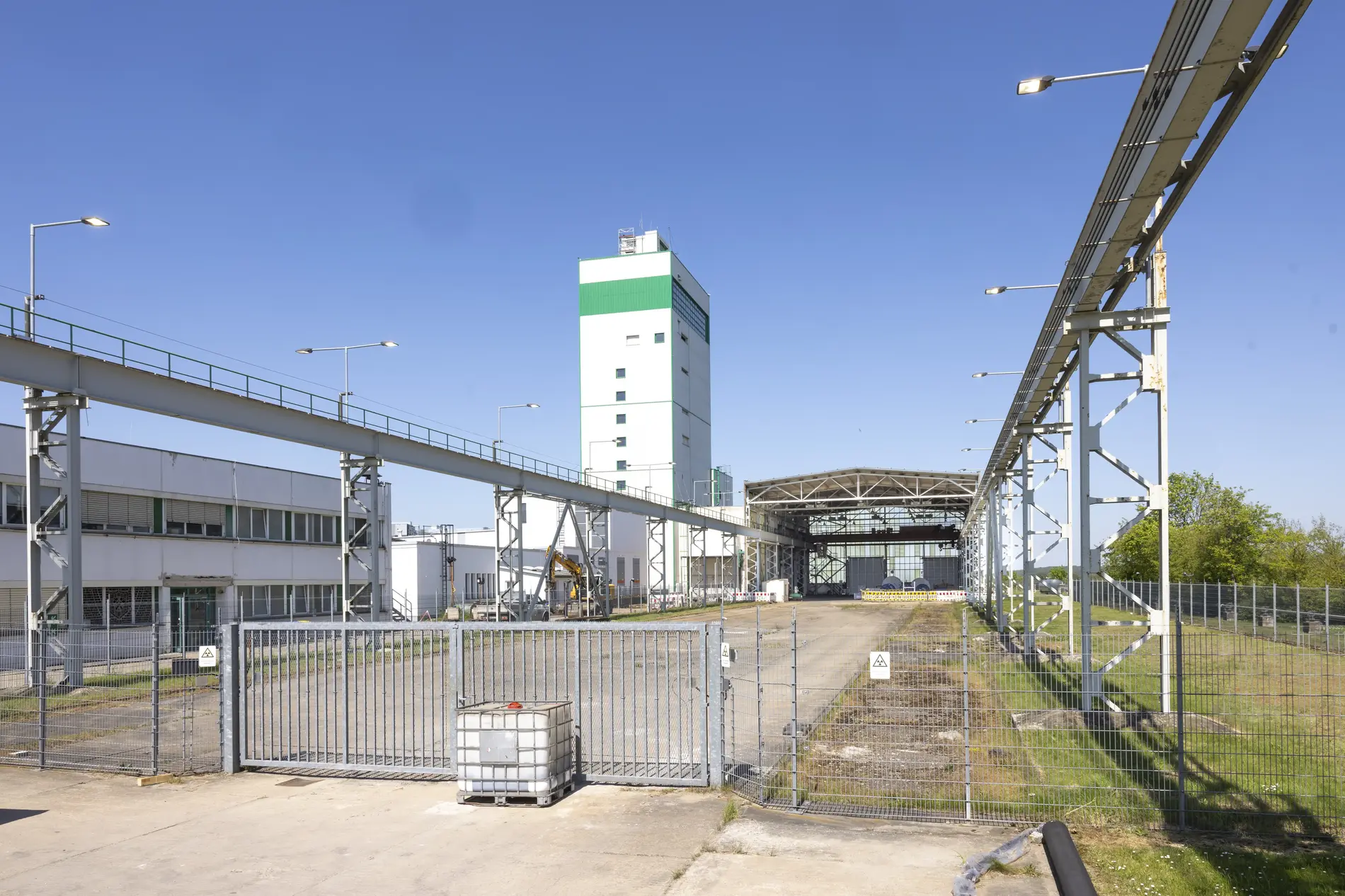 Vor blauem Himmel präsentiert sich das Betriebsgelände des Endlagers Morsleben hinter einem großen Tor. Der weiße Förderturm ragt in die Höhe. 