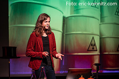 Eine junge Frau mit langen Haaren spricht auf einer Bühne. Im Hintergrund sind die Zeichnungen gelber Atomfässer zu sehen.