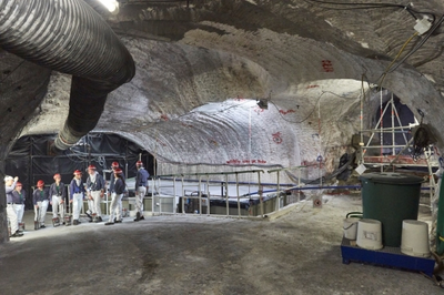 Es ist ein Hohlraum unter Tage zu sehen. In diesem befindet sich ein Speicherbecken, was mit einer Plane abgedeckt ist. Über eine blaue Rohrleitung läuft die Zutrittslösung in das Becken. Vor dem Becken steht eine Gruppe von Besucher*innen, die sich diesen Arbeitsplatz unter Tage anschauen.