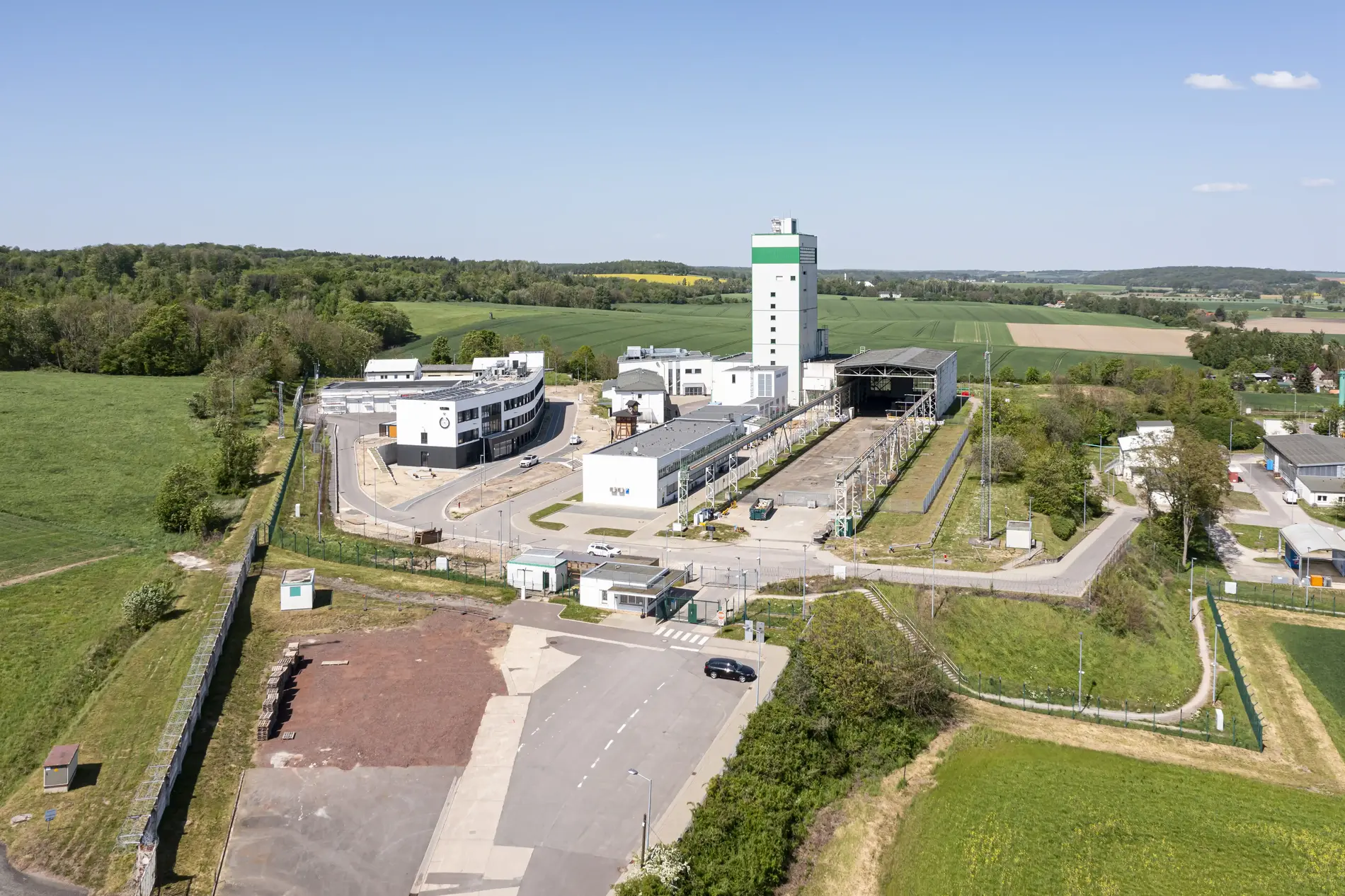 Das Endlager Morsleben fotografiert aus der Luft. Das Betriebsgelände samt weißem Förderturm ist umgeben von grünen Feldern und Wälder bei blauem Himmel.