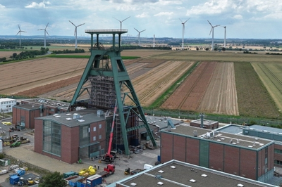 Zu sehen ist ein Förderturm und eine darunterliegende Schachthalle auf einem Gelände mit weiteren Bauten.