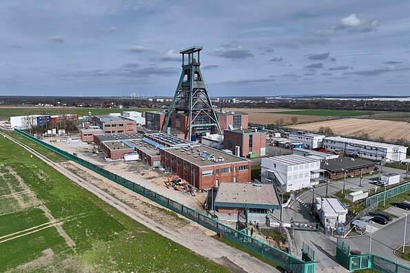 Das Drohnenfoto zeigt eine Industrieanlage mit Gebäuden und einem Förderturm inmitten von grünen Feldern vor einem bewölkten Himmel.