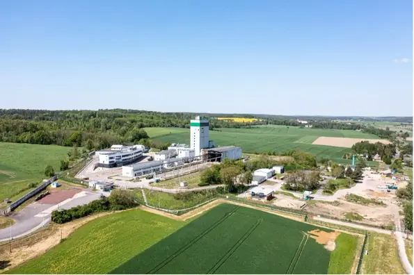 Luftansicht auf eine Industrieanlage inmitten von Feldern und Bäumen. Im Zentrum der Anlage steht ein weißer Turm.