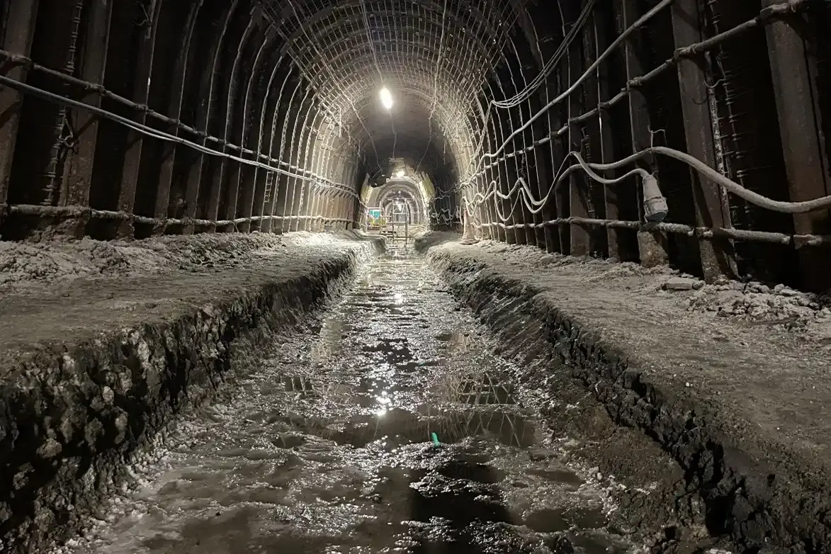 Das Bild zeigt eine Strecke unter Tage. Im Boden verläuft ein Kanal, in dem sich Pfützen befinden. Die komplette Strecke ist mit bogenförmigen Stahlstangen ausgekleidet. 