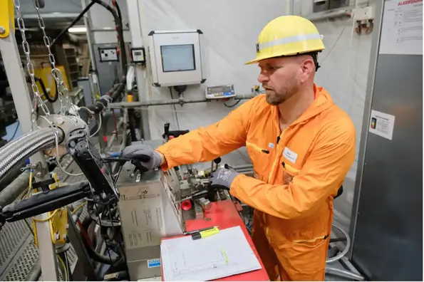 Zu sehen ist ein Mann in orangefarbenem Overall mit gelbem Helm. Er steht vor einem Bedienpult für die Bohranlage.