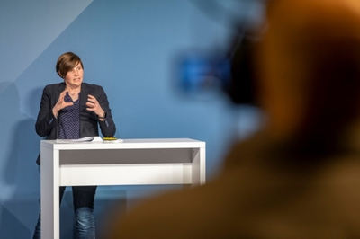 Eine Frau mit dunklen, halblangen Haaren steht an einem weißen Tisch und spricht in eine im Vordergrund schemenhaft erkennbare Kamera.