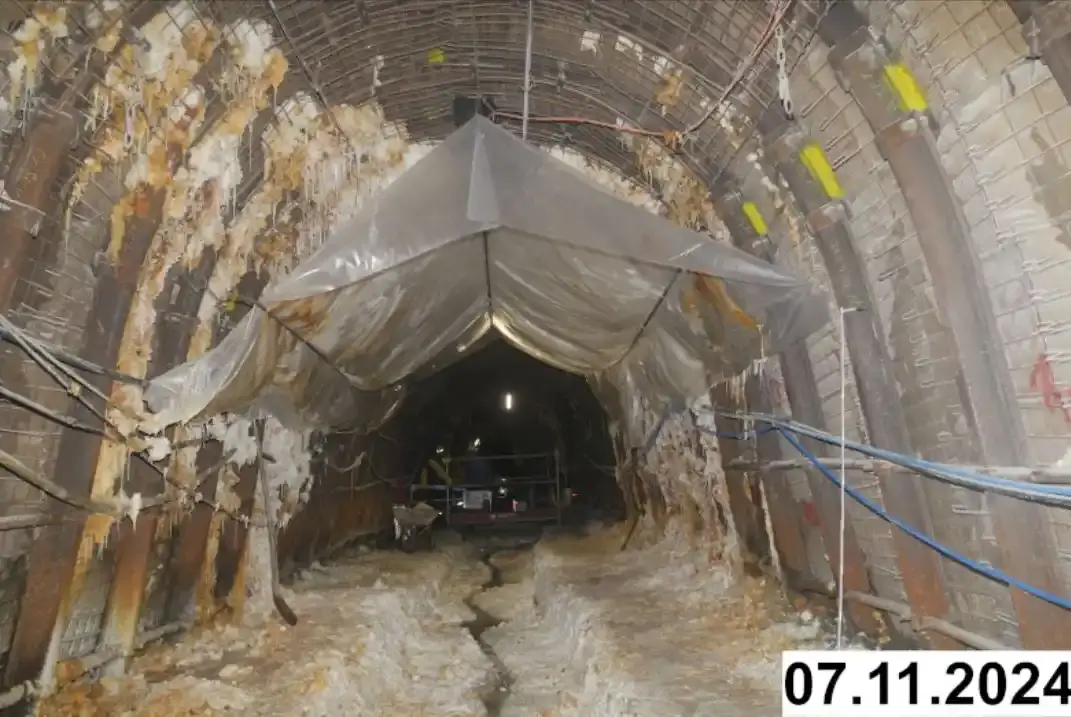 Foto einer bogenförmigen Strecke unter Tage. An den Seiten und der Decke bilden sich weiße Verkrustungen. Eine Plastikplane ist unter der Decke aufgespannt.