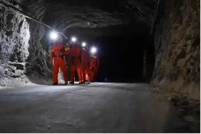 Vier Personen in orangefarbenen Schutzanzügen und Helmlampen gehen hintereinander einen Tunnel in einem Bergwerk entlang.