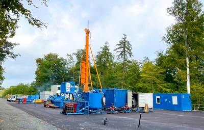 Das Bild zeigt den Bohrplatz zur Erkundungsbohrung Remlingen 18. In der Mitte des Bildes befindet sich ein orangefarbener Bohrturm, umgeben von mehreren blauen Containern. Diese stehen, inmitten eines grünen Waldes, auf einer flach asphaltierten Fläche. Im Hintergrund ist blauer Himmel.