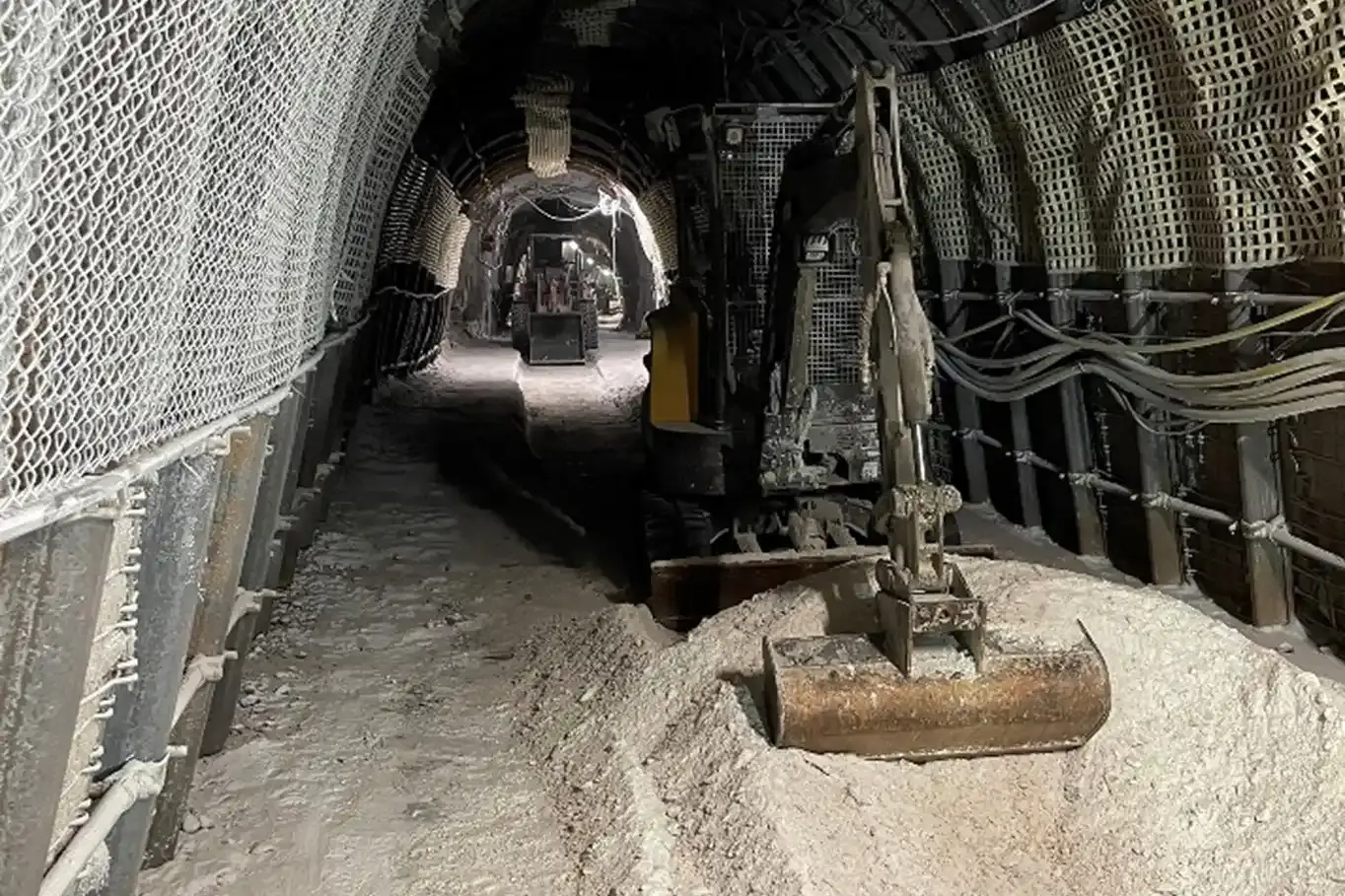 Das Bild zeigt zwei Minibagger unter Tage in einer Bergwerksstrecke. Die Bagger entfernen Teile des Bodens. Die Wände und Decke der Strecke sind mit Bögen aus Stahl und Zaunelementen gesichert. Die Schaufel des vorderen Minibaggers ruht auf einem Haufen Salzgestein. An der rechten Wand führen graue Kabel entlang.