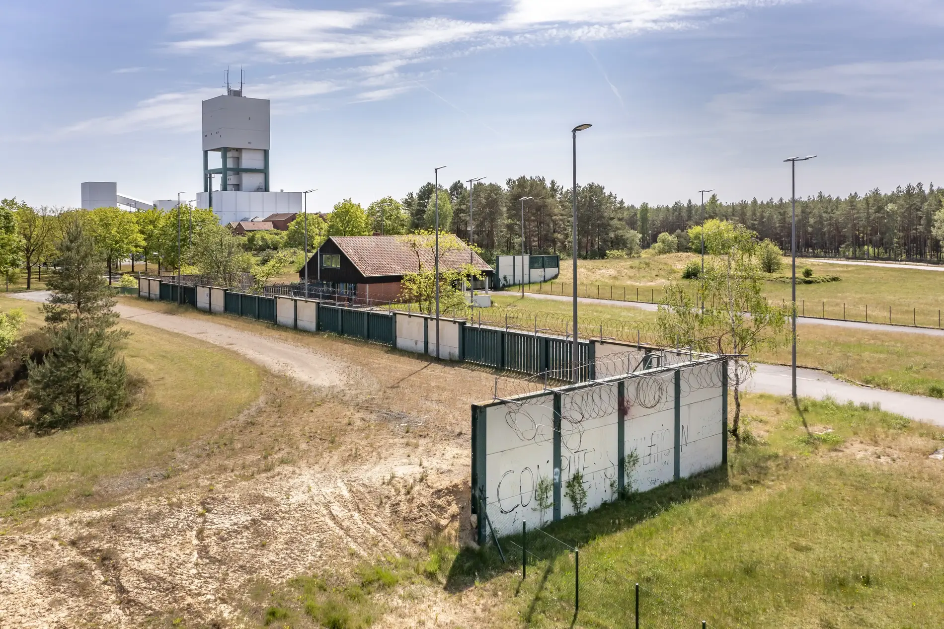 Im Vordergrund sind Reste einer weißen Mauer zu sehen. Im Hintergrund ragt der Förderturm des Bergwerks Gorleben hinter Bäumen in die Höhe. 