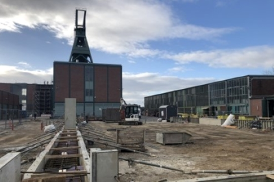 Das Bild zeigt einen Platz auf dem Betriebsgelände von Konrad 1. Im Hintergrund sind der Förderturm und daneben eine große Backsteinhalle zu sehen. Im Vordergrund sind Schalungswände für ein Fundament der temporären Werkstatt zu sehen.