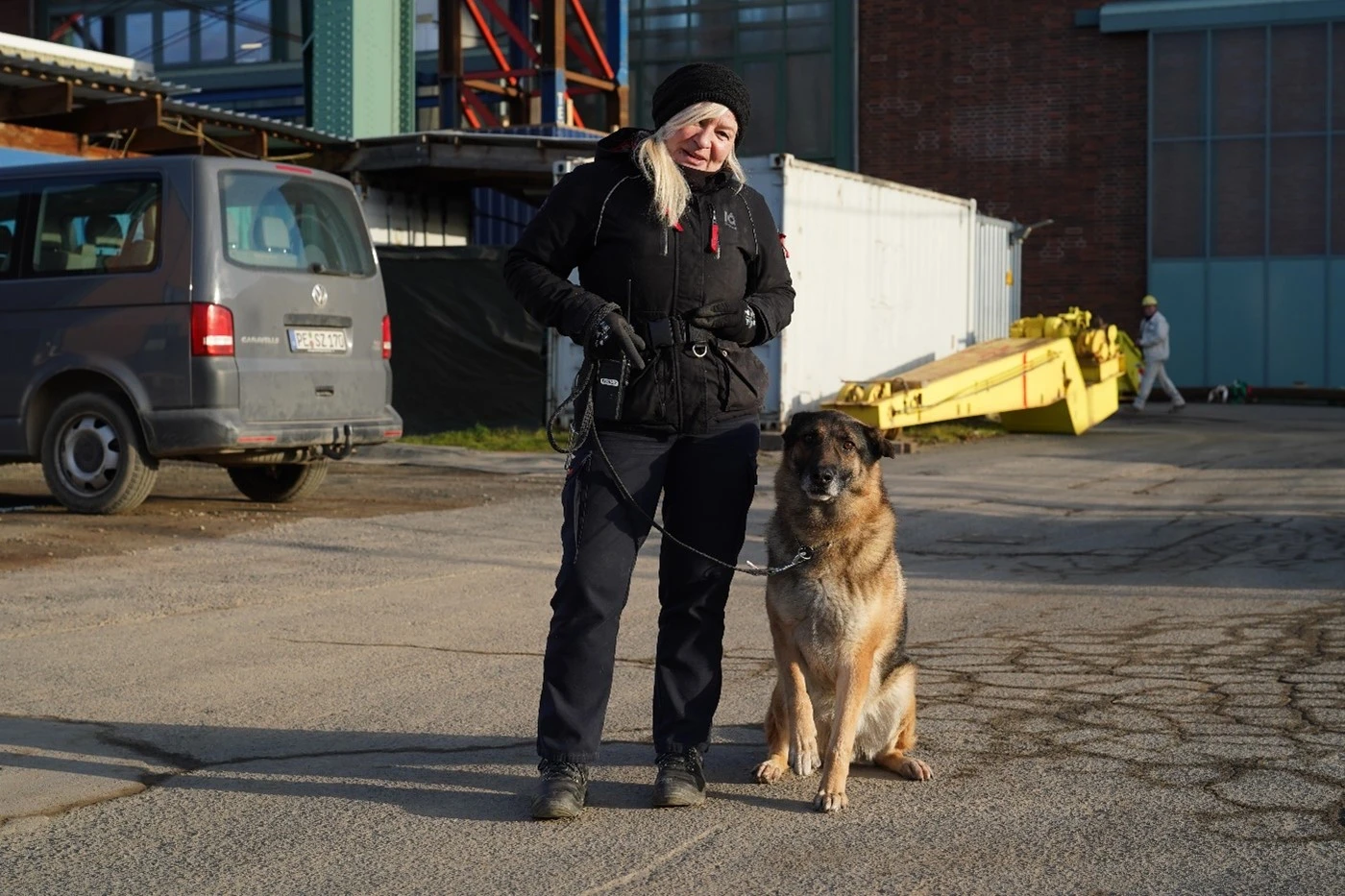 Eine Frau in schwarzer Kleidung steht vor einer Halle. Neben ihr sitzt ein deutscher Schäferhund.