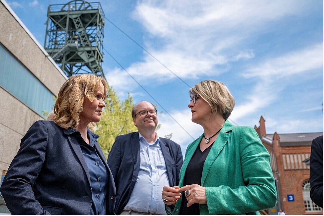 Drei Personen unterhalten sich miteinander. Im Hintergrund befindet sich das Fördergerüst der Schachtanlage Asse II vor einem blauen Himmel mit weißen Schleierwolken.