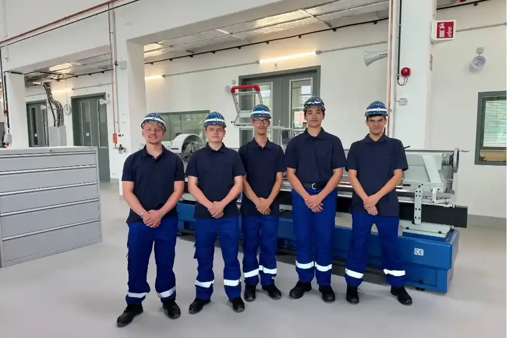 Foto von fünf jungen Männern in blauer Arbeitskleidung in einer Werkstatt. Die Personen tragen schwarze Stiefel und blaue Schutzhelme.