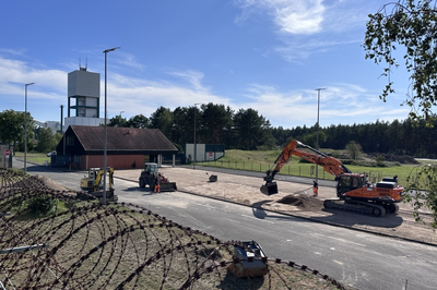 Foto einer Baustelle mit Radlader, Minibagger und orangefarbenem großen Bagger vor einer geglätteten Kiesfläche. Im Hintergrund ist der Förderturm des Bergwerks Gorleben vor einem blauen Himmel mit weißen Wolken zu sehen.