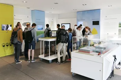 Das Bild zeigt eine Gruppe von Besucher*innen in der Infostelle Konrad. Die Besucher*innen gucken sich die Ausstellung und ein Modell des Bergwerks an.