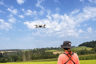 Das Foto zeigt eine Peron mit langen Haaren und Hut von hinten. Die Person steuert eine aufsteigende Drohne über ein Feld bei blauem Himmel.