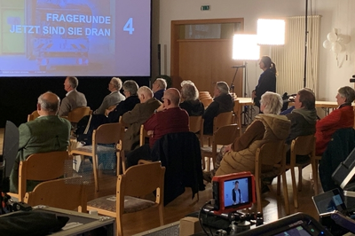 Das Foto zeigt einen Blick in die Veranstaltung. Rechts steht eine Zuschauerin am Mikrofon und stellt eine Frage. Das Publikum schaut auf eine Leinwand mit Präsentation.