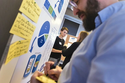 Ein Mann steht vor einer Plakatwand mit Diagrammen und aufgeklebten Post-its. Im Hintergrund sprechen zwei Frauen miteinander.