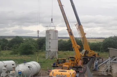 Zwei Kräne stehen vor grüner Landschaft, einer hebt ein Silo an