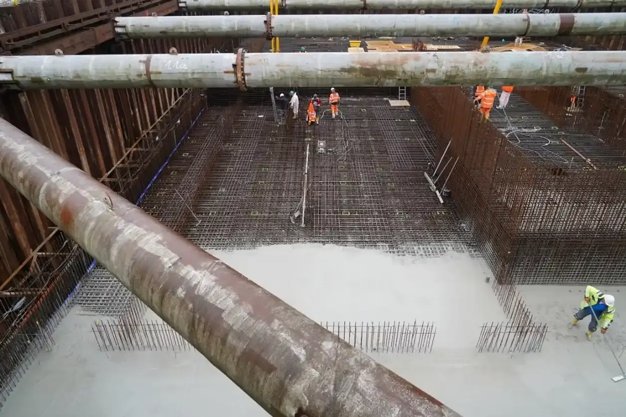 Bauarbeiter führen Betonierarbeiten in einer Baugrube aus. Um sie herum ragen Stahlstäbe aus dem Boden. Die Bauleute bewegen sich über ein Drahtgeflecht, über das nach und nach flüssiger Beton gegossen wird.