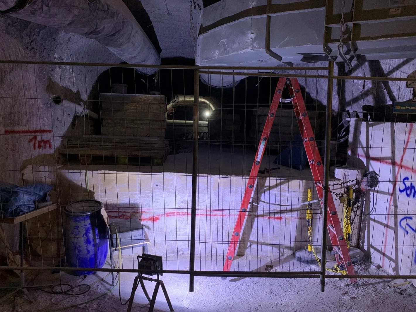 „Bildbeschreibung, Barrierefreiheit“: Hinter einem Bauzaun liegt eine Strecke, deren Boden mit einer 1,20 Meter hohen Schicht aus Spezialbeton bedeckt ist. Die Höhe der Strecke ist dadurch knapp halbiert. An der Decke hängen links und rechts Führungen für Luft und Radongase. 