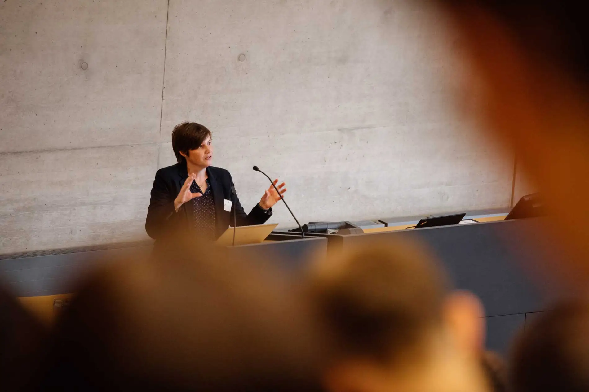 Das Bild zeigt einen Hörsaal, vor dem eine Frau steht und in zwei Mikrofone spricht. Die Frau hat kurze braune Haare und trägt einen schwarzen Blazer, sie gestikuliert. Zuhörende in den Rängen sind unscharf von hinten zu sehen. 