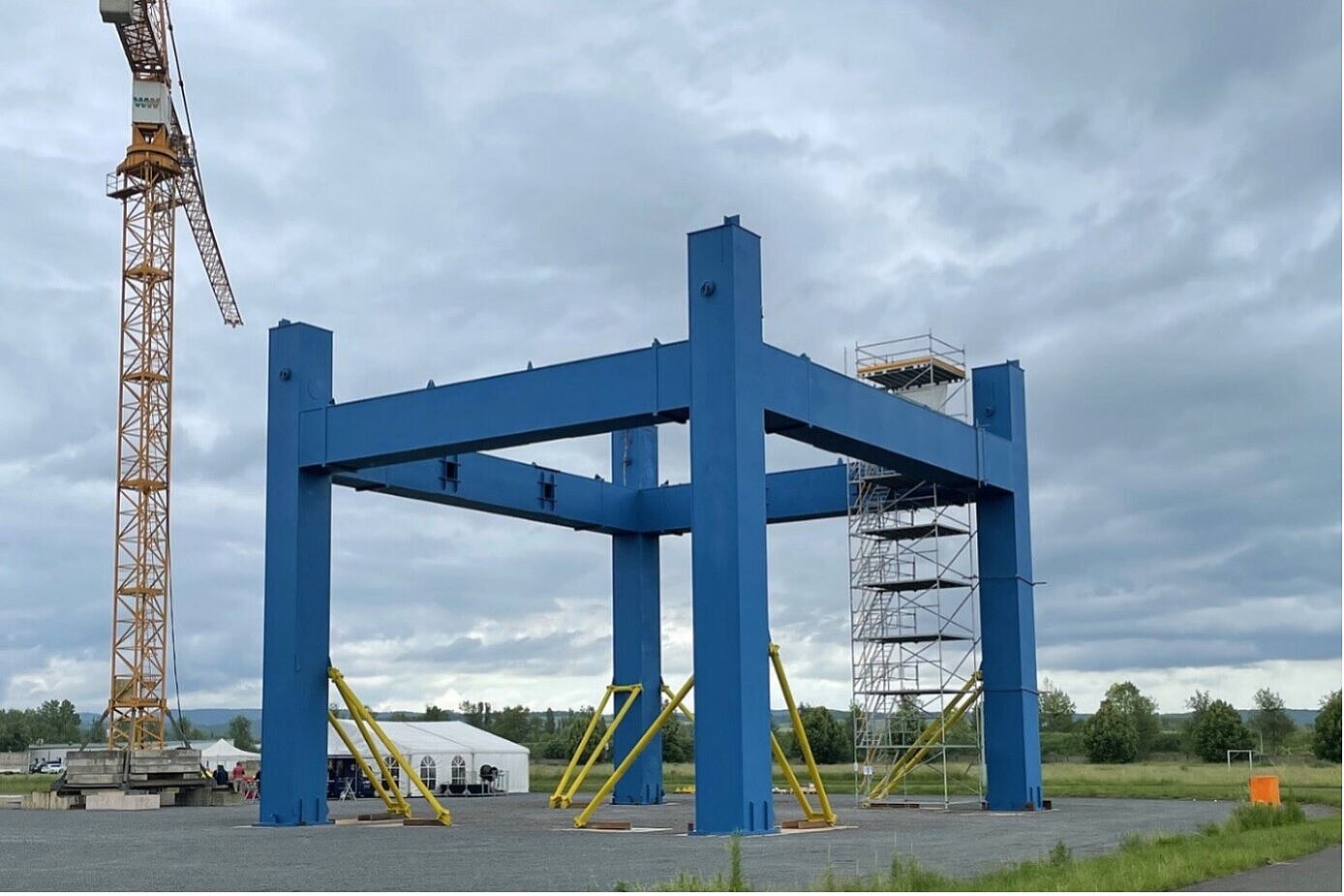  Das Bild zeigt eine große, blaue Stahlkonstruktion auf einer Baustelle im Freien, mit gelben Verstrebungen zur Stabilisierung. Im Hintergrund sind ein Baukran, ein Gerüst, Zelte und eine grüne Landschaft unter einem bewölkten Himmel zu sehen.